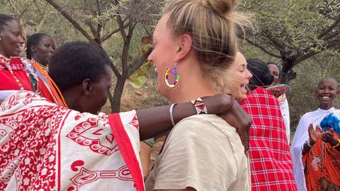       Tourist laughing with Maasai women wearing colourful beads and patterned fabrics beneath thorny trees.
  