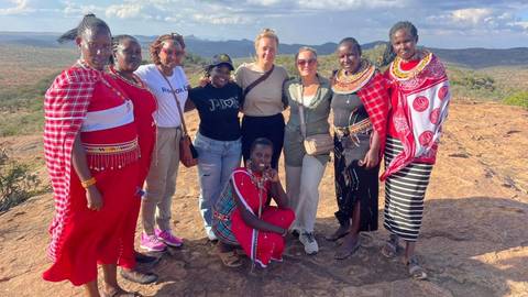       Group photo of travellers and Maasai women standing on rocky viewpoint with expansive savannah backdrop.
  