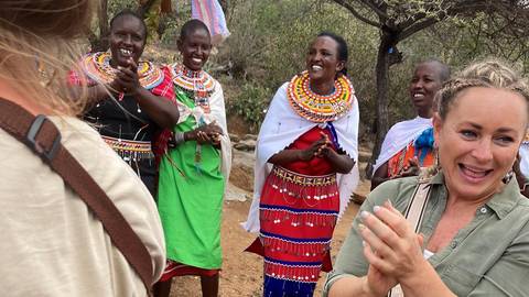       Travellers laughing and clapping alongside Maasai women in colourful traditional dress during cultural visit.
  