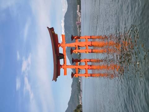       Iconic bright vermilion torii gate of Itsukushima Shrine standing in calm blue sea water with distant mountains and town on the shore.
  