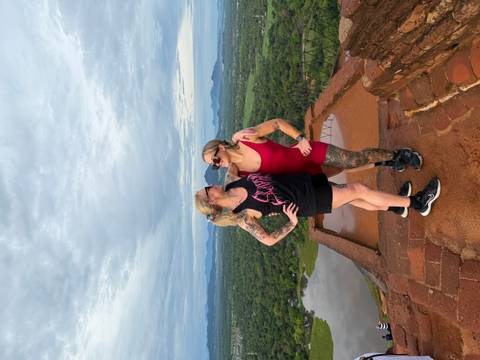       Two friends pose triumphantly on a brick summit with lush forest and Sigiriya Rock in the distance
  