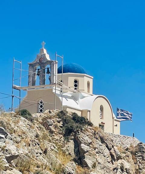       Cream-colored Cycladic church with blue dome and Greek flag set against clear sky, scaffolding on bell tower
  