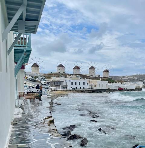       Iconic windmills overlooking Mykonos town with waves splashing along the shore
  