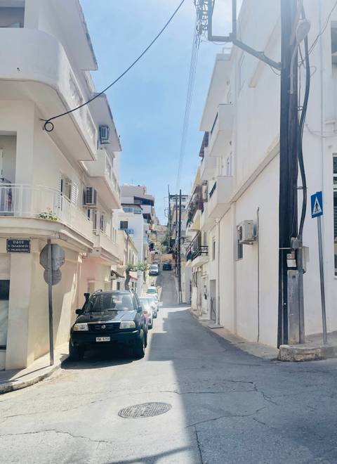       Steep urban street lined with compact white buildings and parked cars on a Greek island
  
