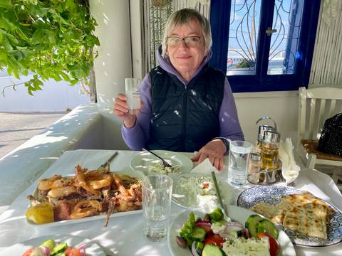       Smiling older woman enjoying a seafood feast at an outdoor Greek taverna with white walls and a blue-framed window.
  