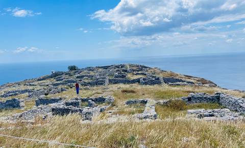       Stone ruins of an ancient hilltop settlement overlooking the deep-blue Aegean Sea with a single visitor wandering among the walls.
  