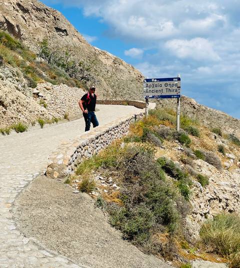       Traveler walking up a stone path past a bilingual road sign for Ancient Thira on a scrubby hillside.
  