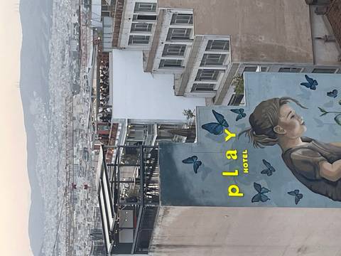       Urban evening scene with a mural of a girl and butterflies on a building facade reading "play hotel" overlooking a sprawling city.
  