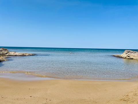       Quiet sandy cove with gentle turquoise water framed by rocky outcrops under a clear blue sky.
  