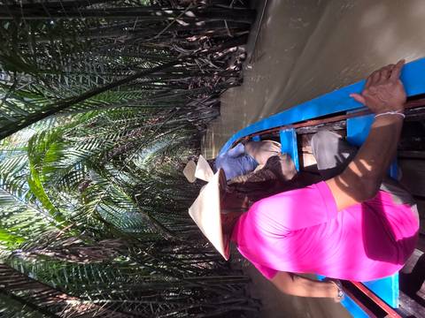       Travelers in a narrow wooden boat glide through dense palm fronds on a muddy Mekong canal.
  