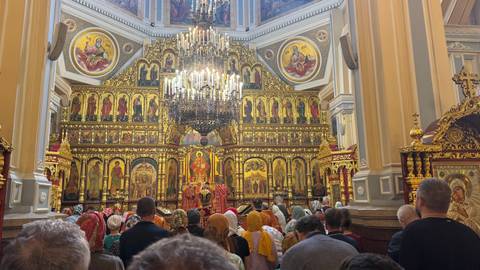       Lavish icon-covered interior of an Orthodox church packed with worshippers under a glittering chandelier.
  