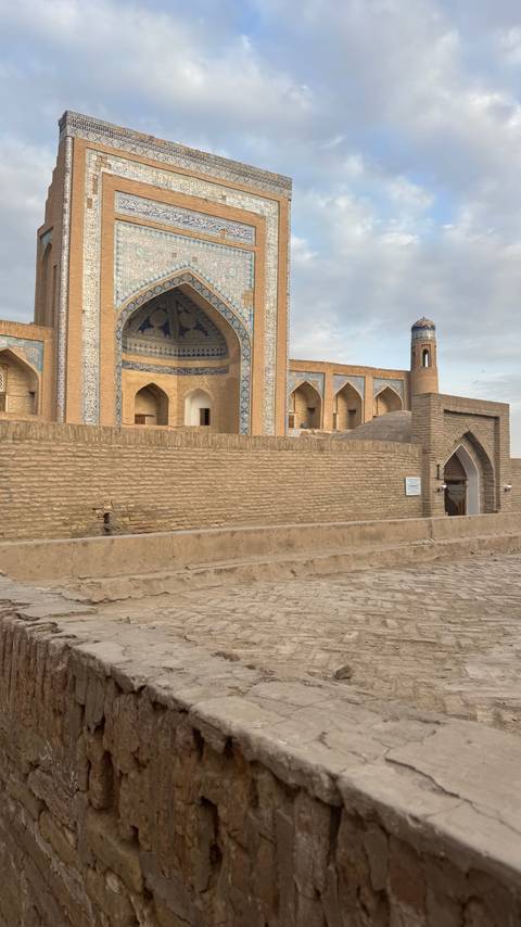       Brick ramparts and arched entrance of a historic madrasa decorated with blue patterned tiles.
  