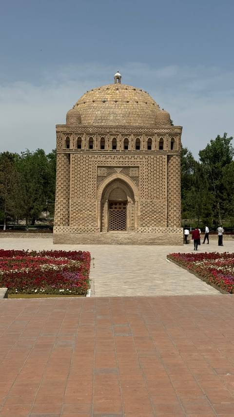      Square patterned brick mausoleum surrounded by landscaped flowerbeds and visitors in the foreground.
  