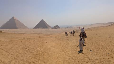       Camel riders approach the Pyramids of Giza across a vast sandy plateau under hazy skies.
  
