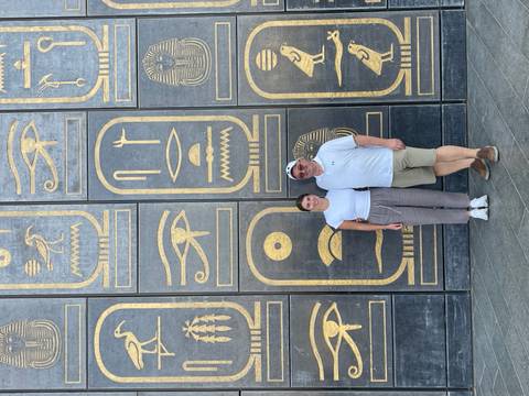       Couple stands before a modern wall decorated with oversized gilt hieroglyphic cartouches.
  