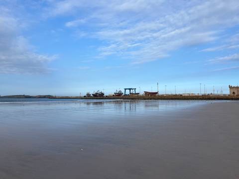       Calm shoreline at low tide facing a small harbour and boatyard under blue skies
  