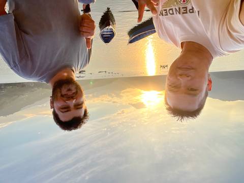       Two friends taking a sunset selfie beside a tranquil lake with mountains in the background.
  