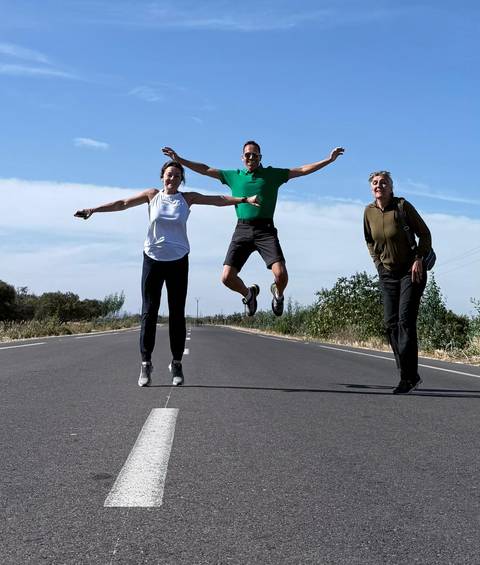       Three travellers strike playful poses, one mid-air jump, on an empty country road under a bright Moroccan sky.
  