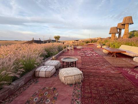       Stylish desert camp terrace decorated with patterned rugs, poufs and lanterns amid waving grass at dawn.
  