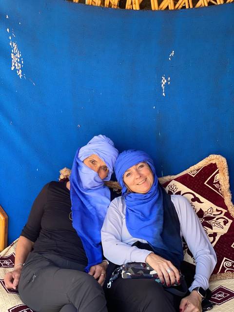       Two women wearing bright blue Tuareg scarves relax on a patterned cushion against a cobalt backdrop.
  