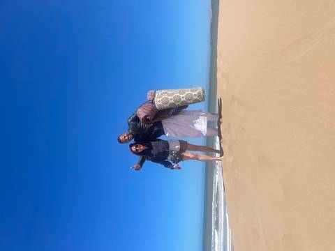      Two people pose happily on a windswept sandy beach dune beneath a cloudless blue sky.
  
