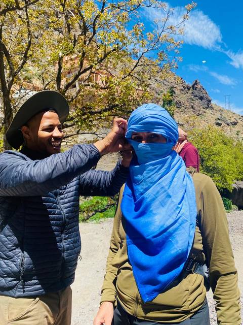       Guide helps a traveller wrap a bright blue headscarf in a mountain village setting.
  