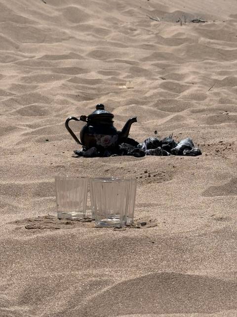       Traditional metal teapot and two glass cups rest on desert sand beside charcoal embers.
  