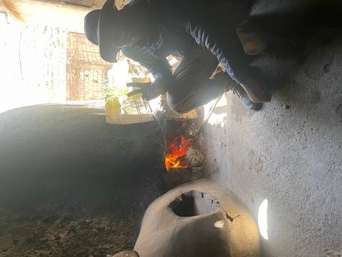       Man sitting beside a traditional clay oven with an open wood fire, flashing a peace sign in dim light.
  