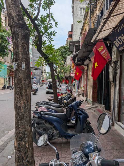       Narrow old-town Hanoi street with motorbikes, shops and national flags.
  