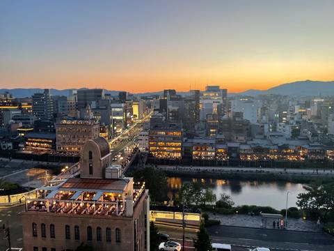       Sunset over Kyoto skyline with glowing orange sky reflecting on the Kamo River and city lights coming alive
  