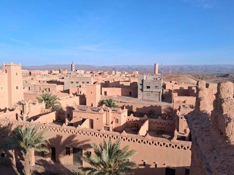       Adobe-colored kasbahs and palm trees of a desert town stretch toward barren Atlas foothills under clear skies.
  