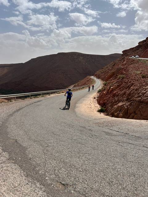       Cyclists riding up a winding mountain road with reddish rocky slopes under a hazy sky.
  