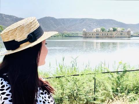       A woman in a straw hat looks across a tranquil lake toward the Jal Mahal water palace backed by hills.
  