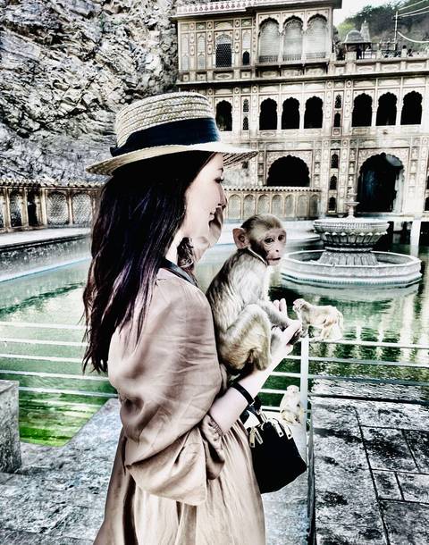       A traveller gently holds a small monkey beside an ornate pool and arched temple walls.
  