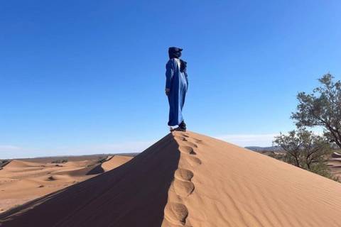       Solo traveler in blue traditional attire standing atop a sharp sand dune ridge under a clear desert sky
  