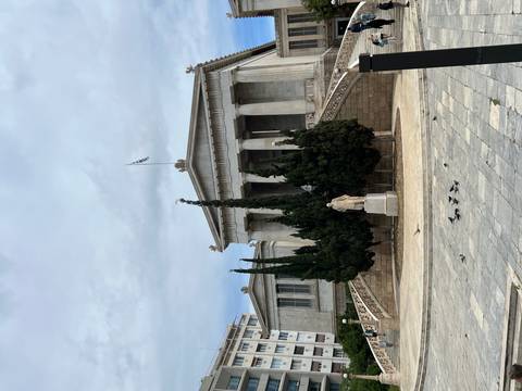       Neoclassical marble building with columned portico and staircases framed by tall cypress trees under a cloudy sky.
  