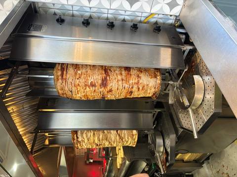       Vertical roasting spits stacked with layers of seasoned meat cook beside shiny stainless steel grills.
  