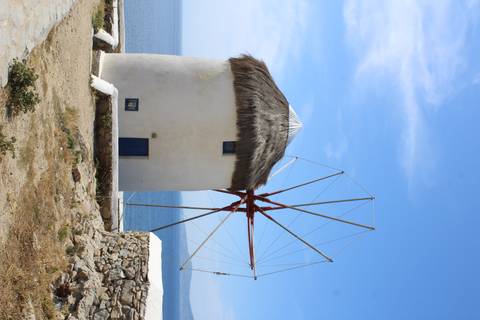       A traditional whitewashed Cycladic windmill with thatched roof stands on a hillside overlooking the Aegean Sea.
  