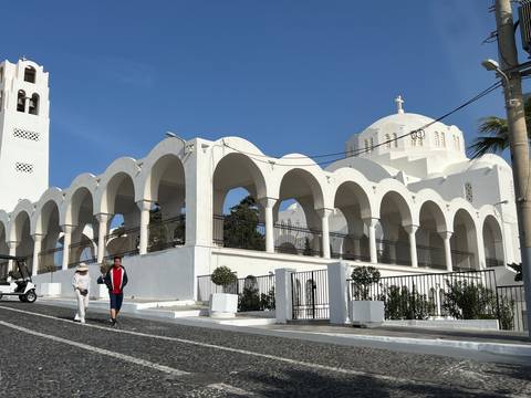       Two people walk past a large white domed church with multiple arches on a sunny island street.
  