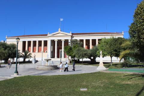       A neoclassical university building with red columns fronts a fountain and green lawn where people stroll.
  