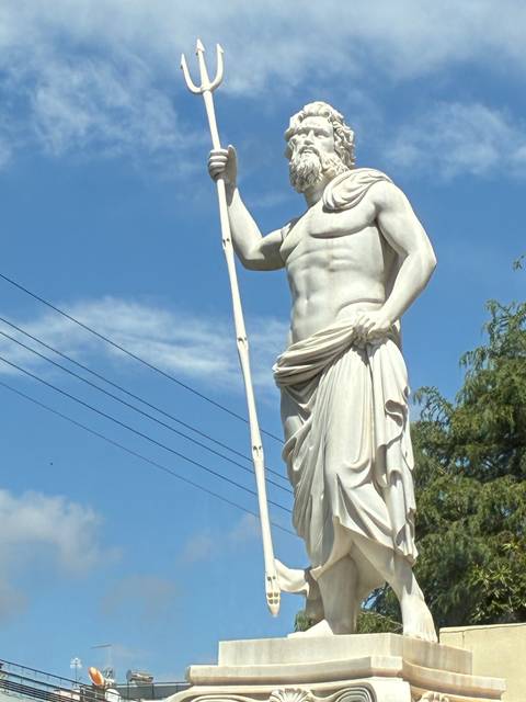       Close-up of a white marble statue of a muscular bearded figure holding a staff against a blue sky.
  