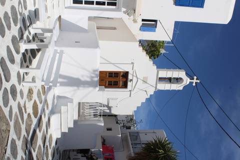       Small white Cycladic chapel with blue shutters and stone courtyard sits beneath overhead wires against a deep blue sky.
  