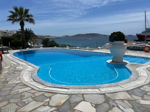       A turquoise hotel infinity pool overlooks the Aegean Sea and distant rocky islets on a bright day.
  