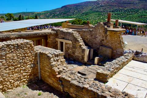       Stone ruins and reconstructed walls of the ancient Palace of Knossos sprawl across a sunny hillside as visitors explore.
  