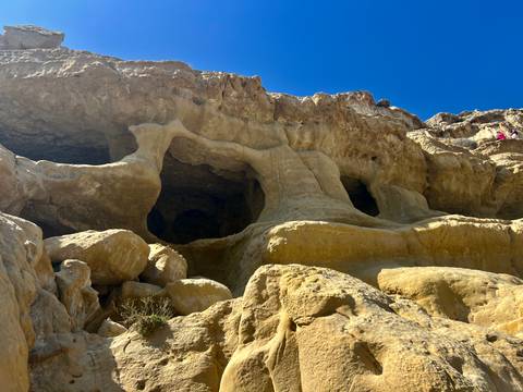       Weathered sandstone cliffs riddled with caves rise steeply against a clear blue sky; two hikers stand near the top.
  