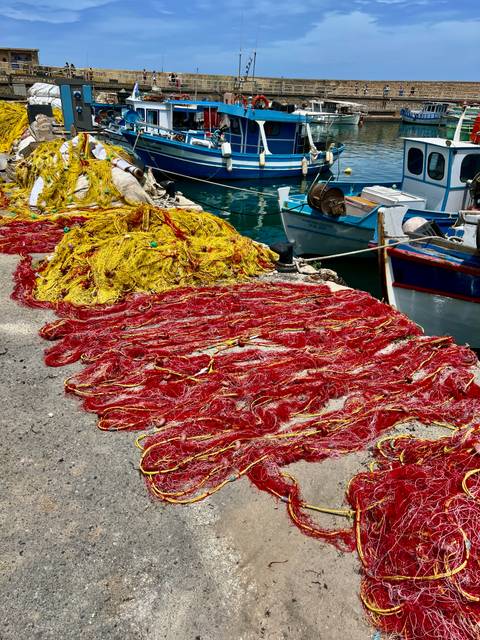       Bright red and yellow fishing nets are piled on a concrete quay beside small blue fishing boats.
  