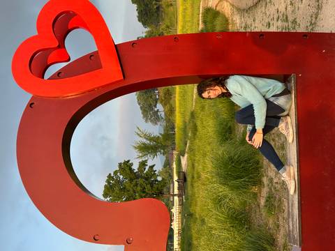       Traveller sitting inside a giant red heart sculpture in a grassy park at sunset.
  