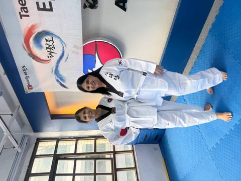       Two women wearing white taekwondo uniforms smile on a blue mat inside a martial arts studio.
  
