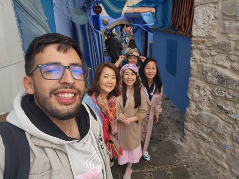       Group smiles for a wider selfie in Chefchaouen's stone alley lined with blue walls and fabrics.
  
