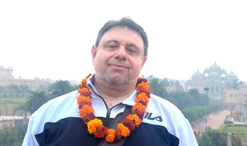       Man wearing marigold garland smiles with hazy temple complex in the misty background.
  
